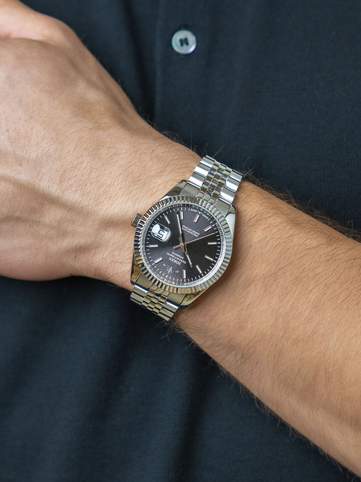 Man wearing the SOREN Momentis Swiss automatic watch, black dial on a 5-link steel bracelet, with hand resting on his chest